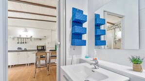 A modern kitchen with white cabinets, a dining area with a chair, and a bathroom with a white sink and blue storage shelves.