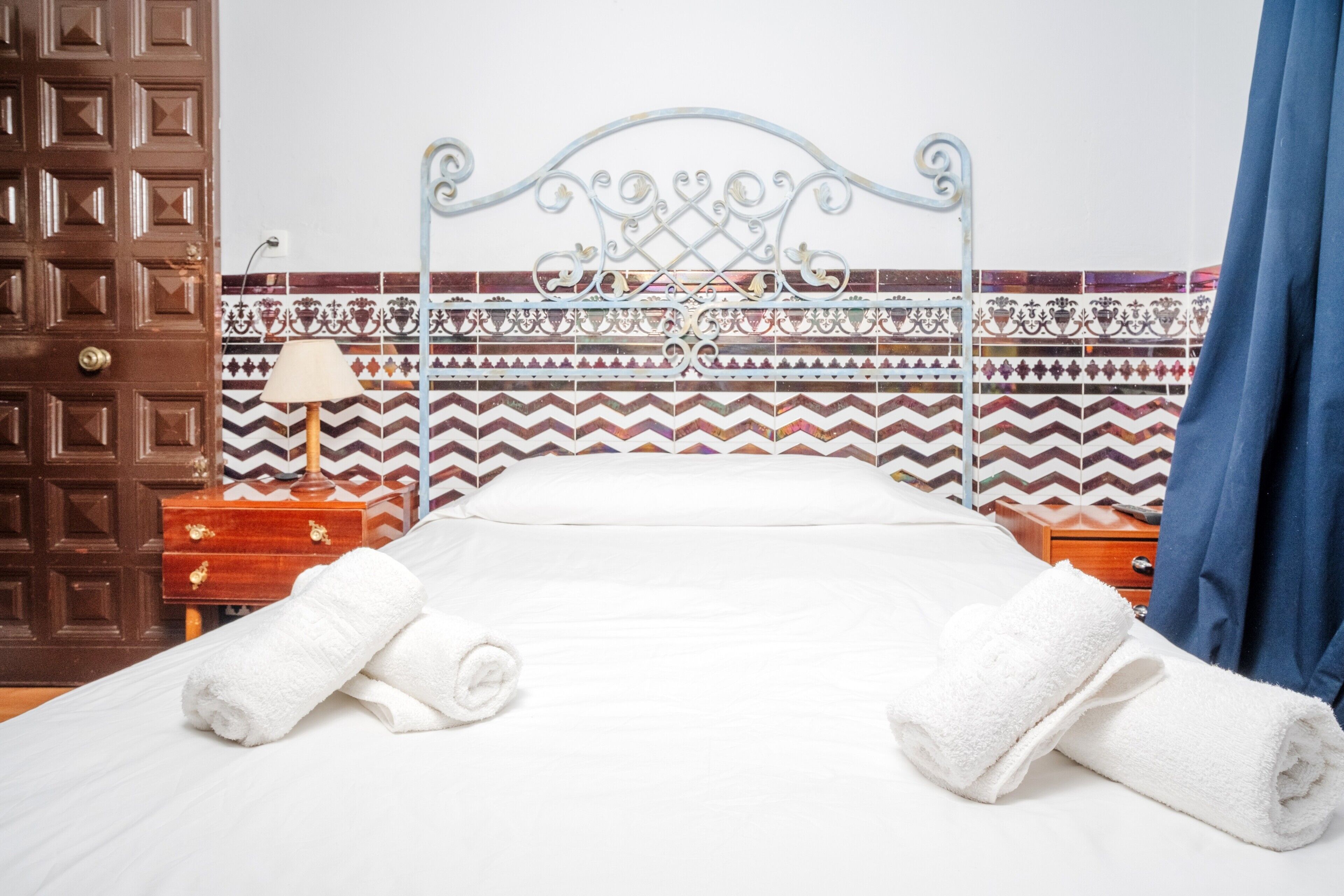 A neatly made bed with white linens and two rolled towels, a wooden nightstand with a lamp, and a decorative metal headboard.