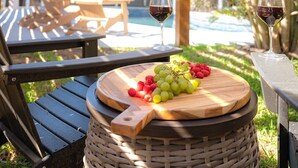 Eine Hollaube mit Tisch und Stühlen darunter, ein rundes Holzschneidebrett mit Trauben und Himbeeren sowie zwei Weingläsern.