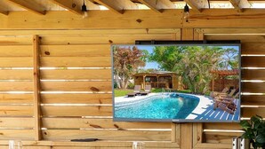 Ein Holzinnenraum mit einem Fernseher, der eine Poolszene zeigt, Holztische mit weißem Geschirr und einer Decke mit freiliegenden Balken.