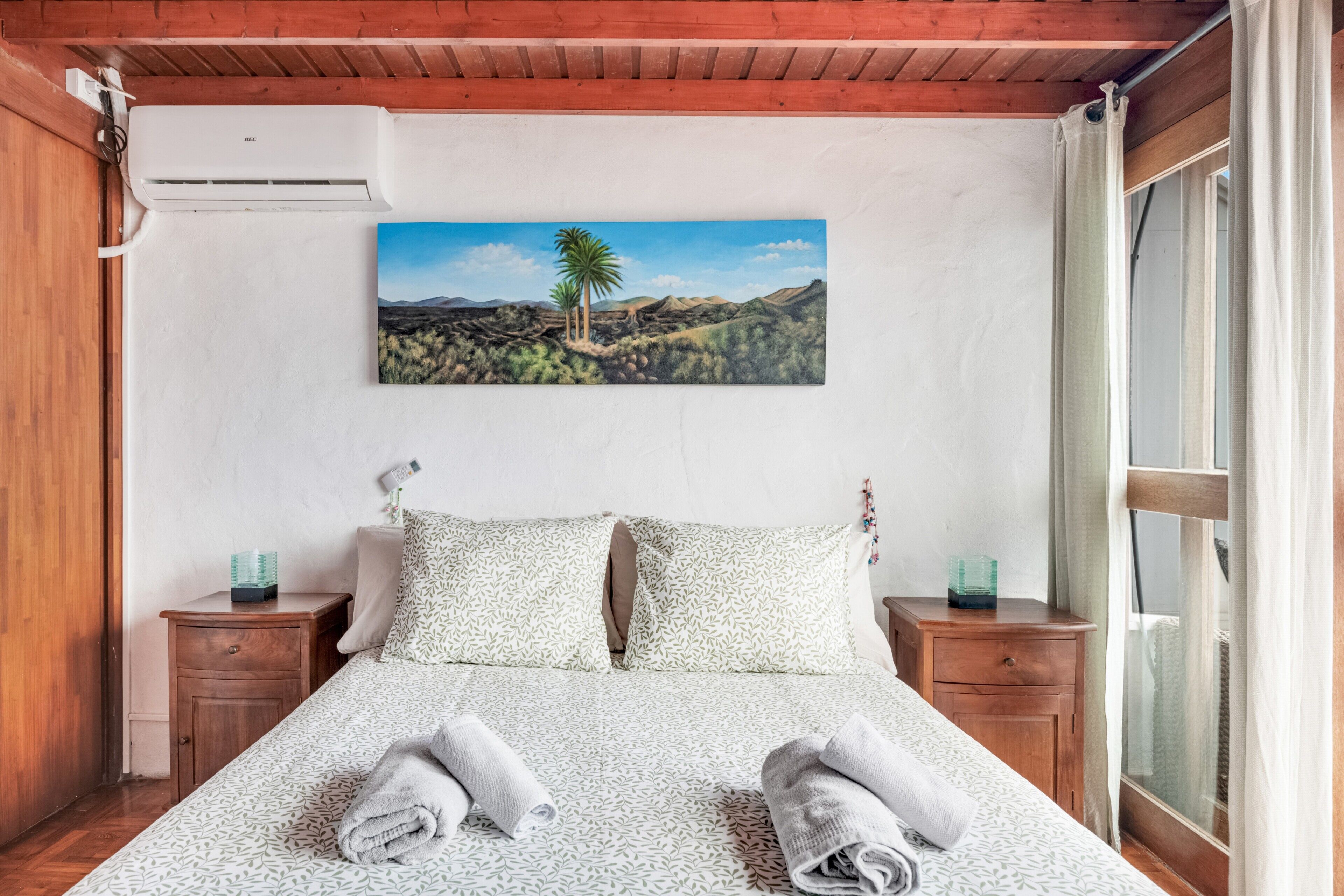 A bedroom with a bed, pillows, a nightstand, a painting, and a window.