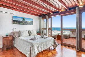 A bedroom with a large bed, wooden ceiling, a painting of a landscape, and a view of the sea through a glass door.