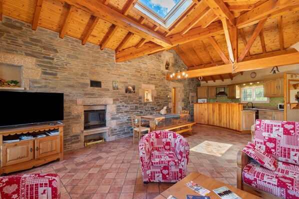A rustic interior with a stone fireplace, wooden beams, a television, and a dining area.