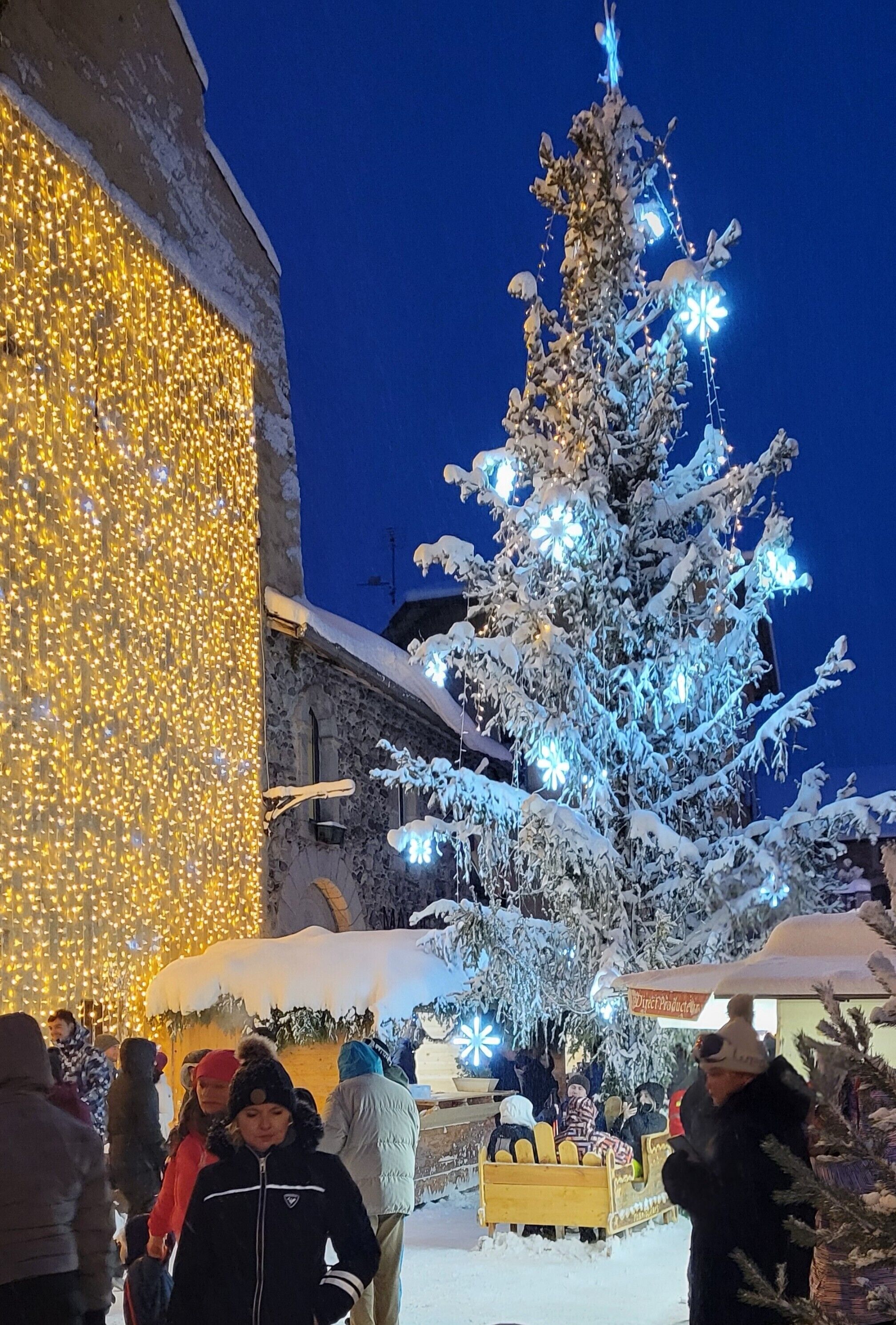 Ein mit Schnee bedeckter Weihnachtsbaum mit Lichterketten, ein Gebäude mit beleuchteten Wänden und eine Menschenmenge.