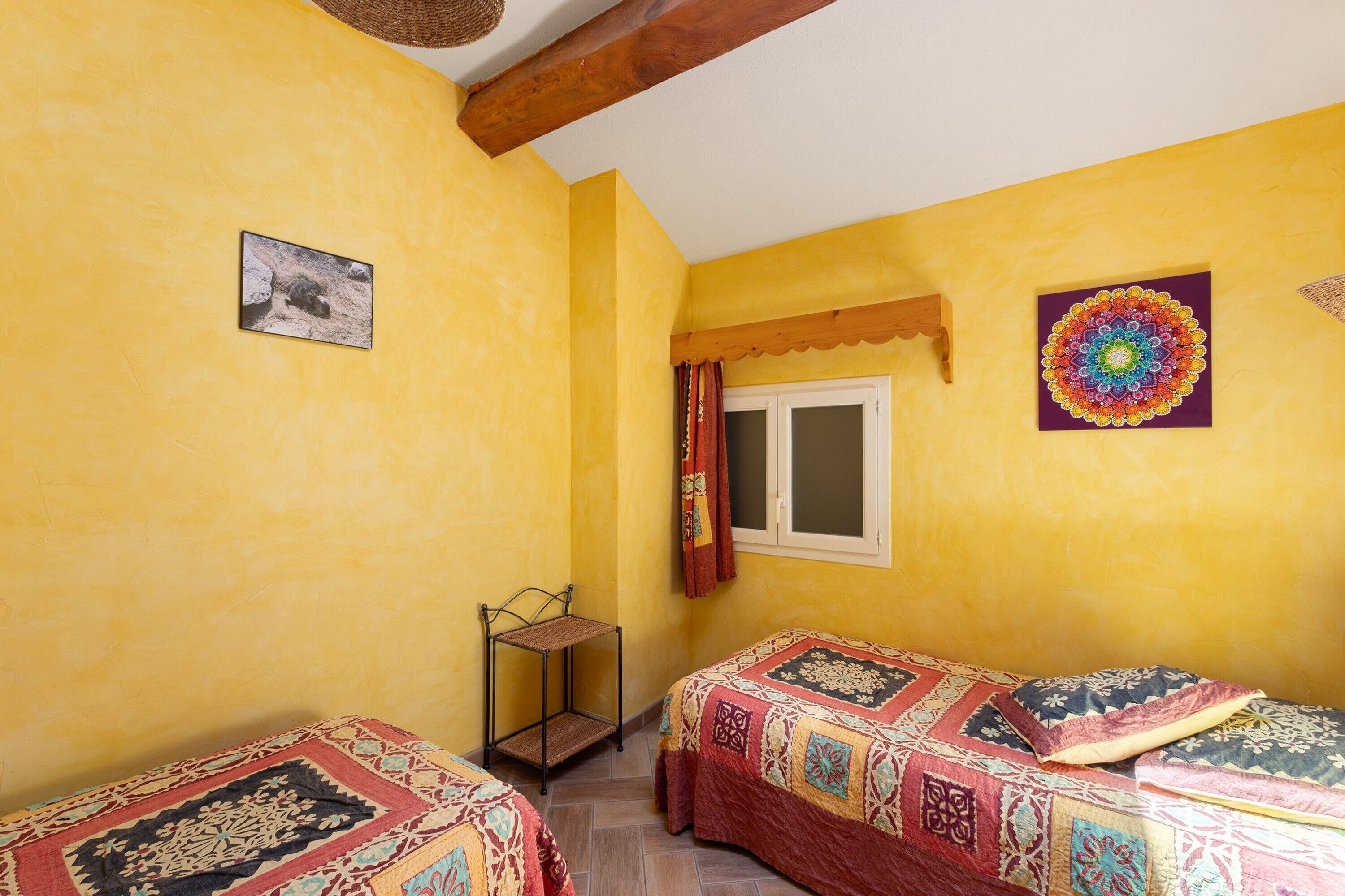 A room with two beds, a window with curtains, a picture on the wall, and a decorative mandala.