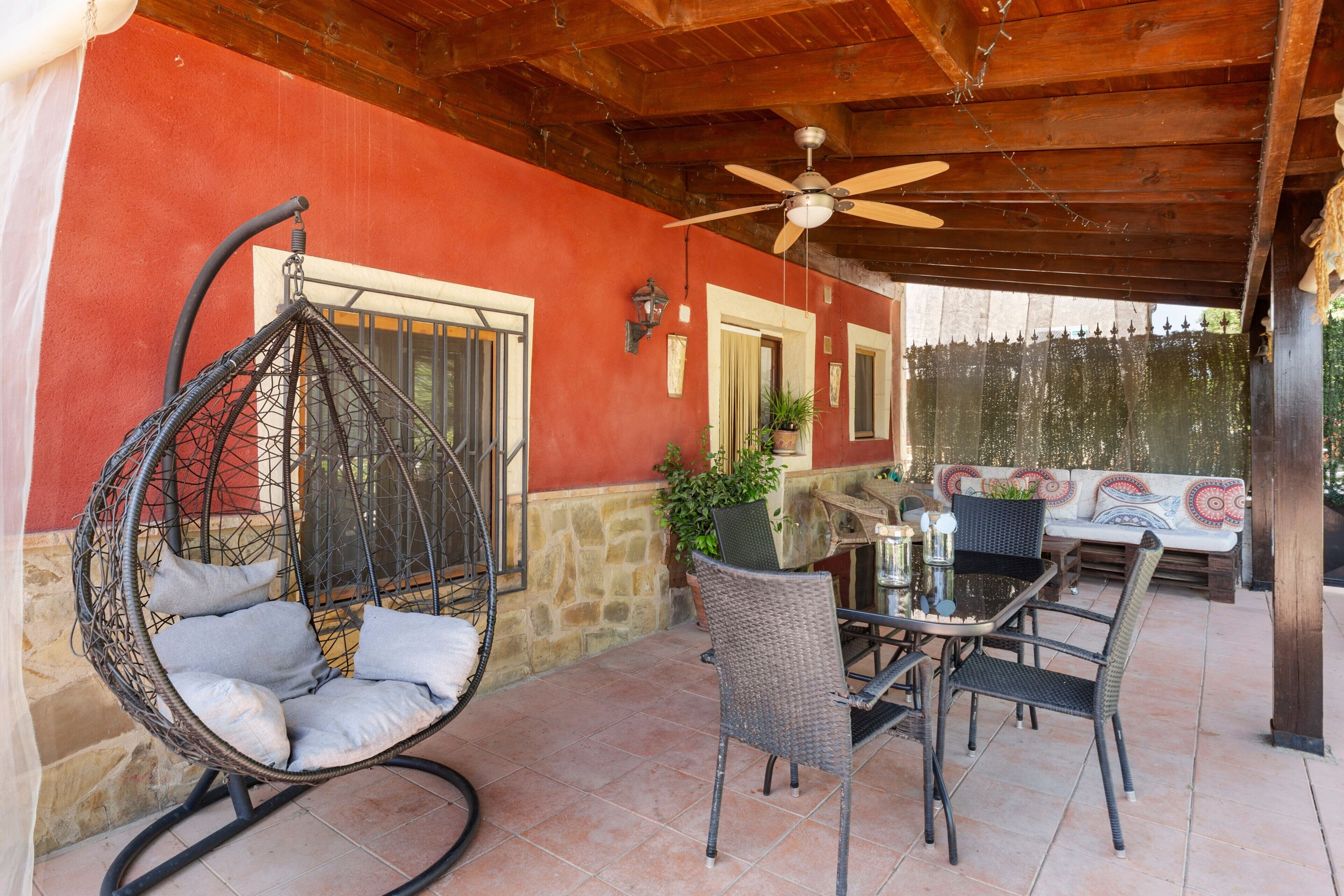 Un patio con una mecedora colgante, una mesa con sillas, un ventilador de techo y un muro de piedra.
