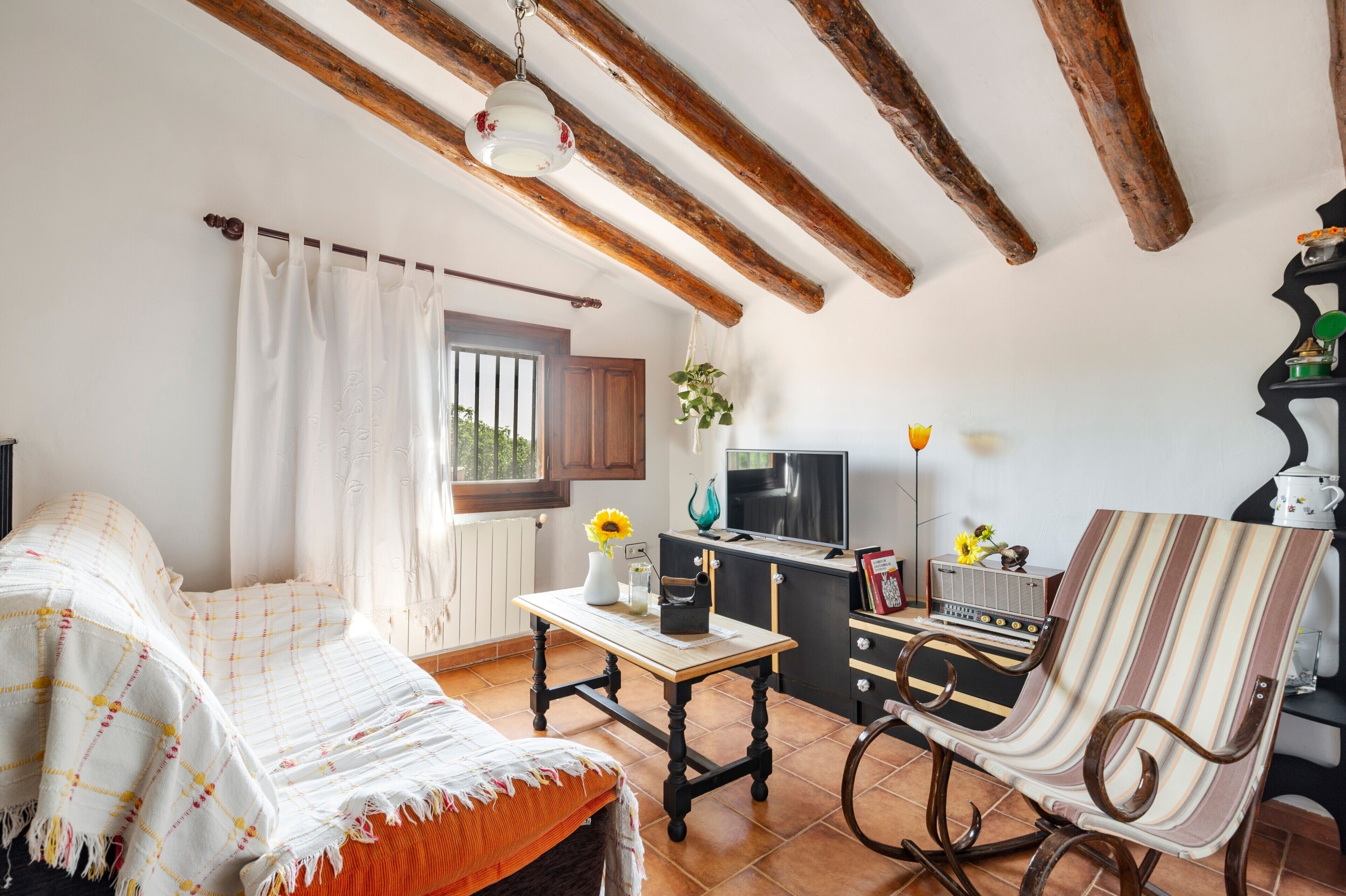 A cozy room with a bed, a table with a vase and a book, a chair, a television, and a kitchen area with a stove and a radio.