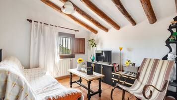 A cozy room with a bed, a table with a vase and a book, a chair, a television, and a kitchen area with a stove and a radio.