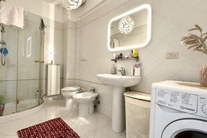 A modern bathroom with a glass-enclosed shower, a white sink, a bidet, and a washing machine. - P034 - Porto Recanati, new three-room apartment with garden (Porto Recanati)