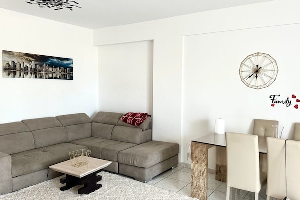 A living room with a sectional sofa, a coffee table, a dining area with chairs, a wall clock, and a cityscape painting.
