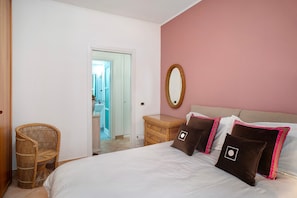 Un dormitorio con una cama, una silla de mimbre, un tocador y un espejo en la pared.