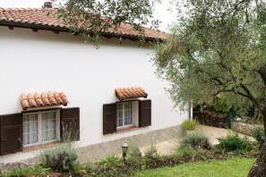 Un bâtiment doté d’un toit en tuiles, de deux fenêtres avec volets et d’un jardin où poussent différentes espèces de plantes.