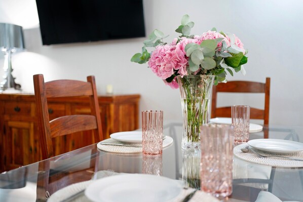Una mesa de comedor con cubierta de vidrio transparente, cristalería rosa y un florero con flores rosadas.