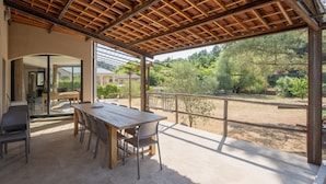 A covered patio with a wooden table and chairs, a glass door leading to an interior space, and a view of a lush garden. - Villa "Luminières I" with Mountain View, Private Pool & Wi-Fi (Les Salelles)