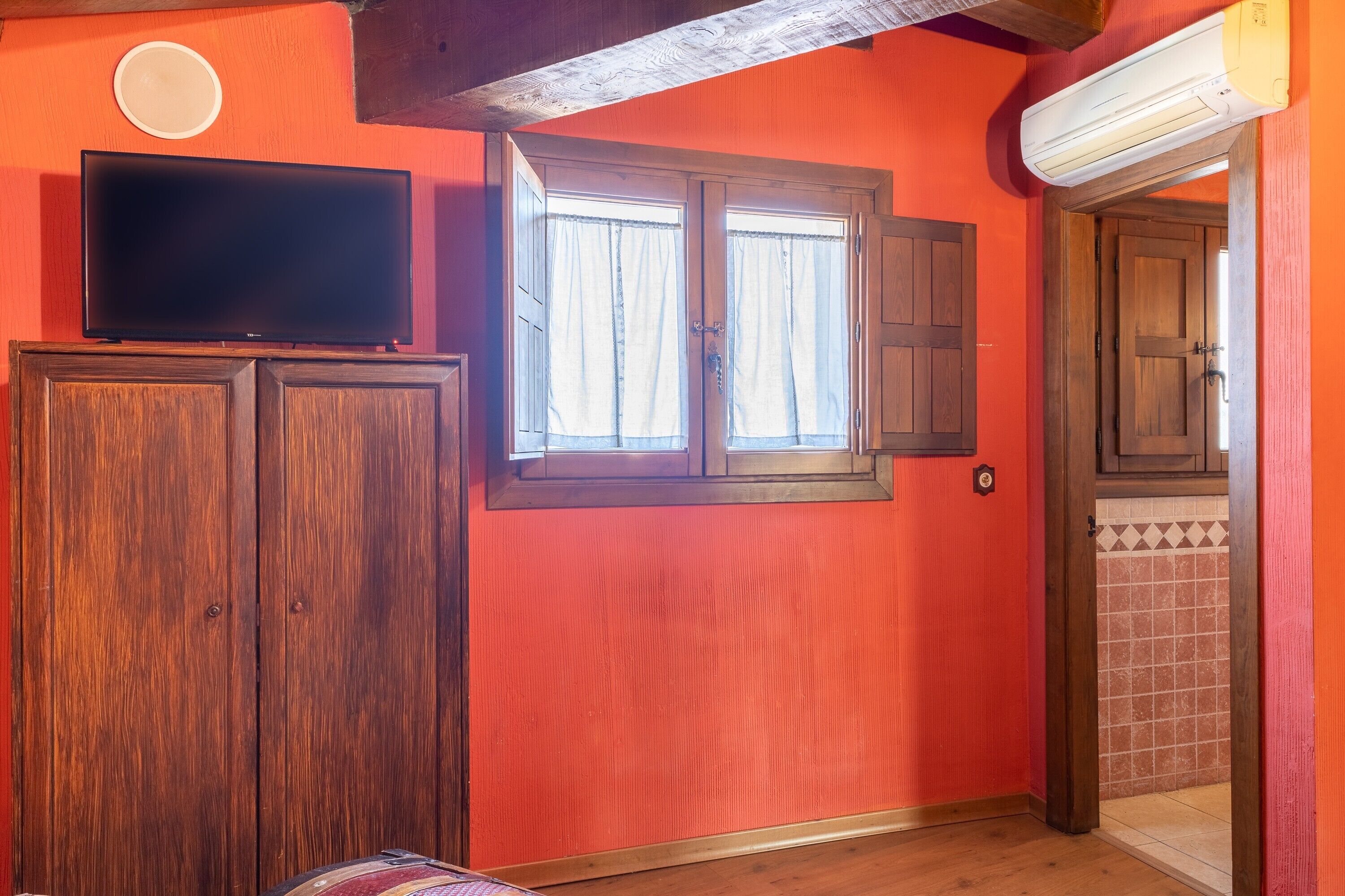 Una habitación con una televisión sobre un mueble de madera, una ventana con cortinas y una puerta que da a otra habitación.