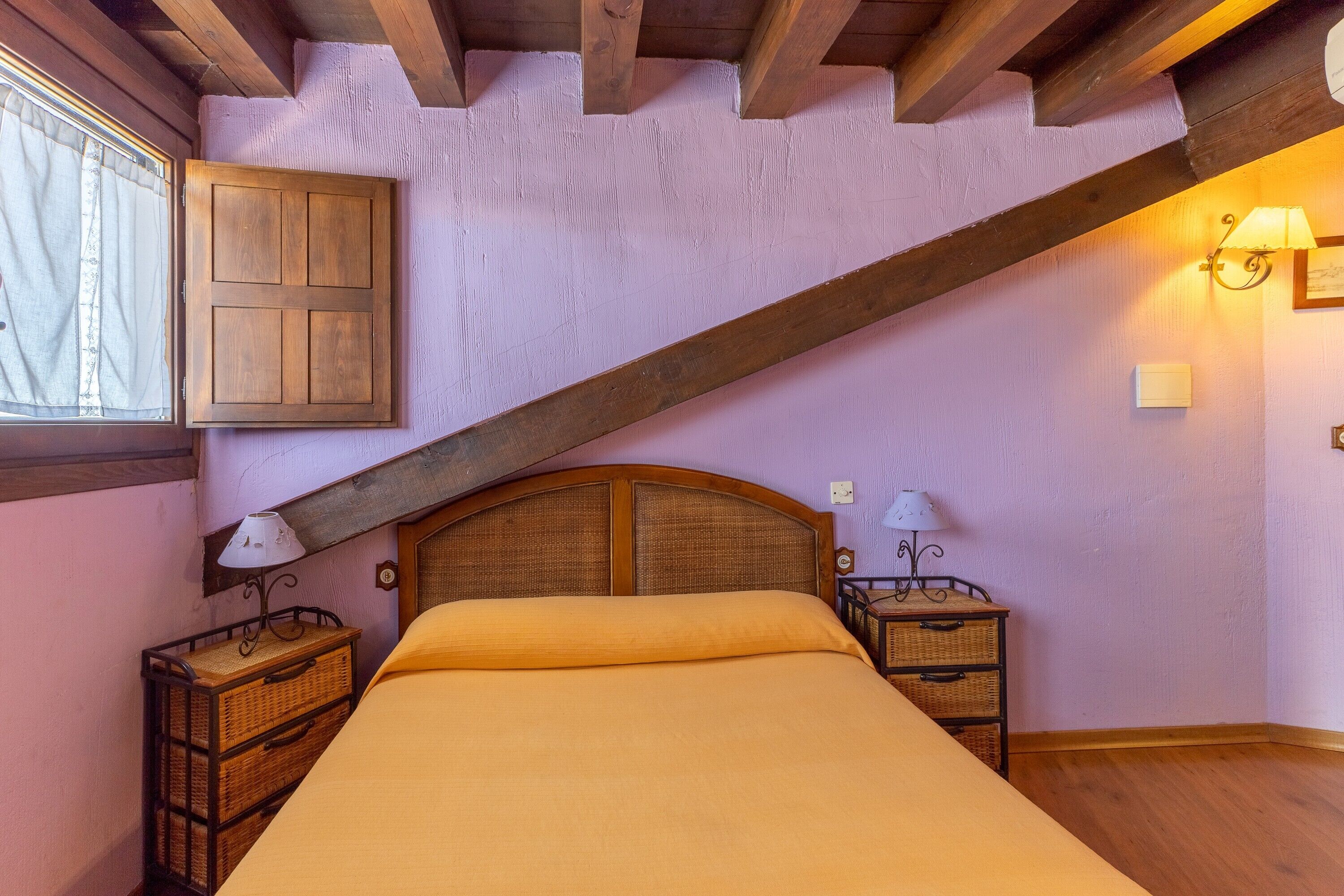 Camera da letto con trave in legno al soffitto, un letto con testiera, due comodini con lampade e una finestra con tende.