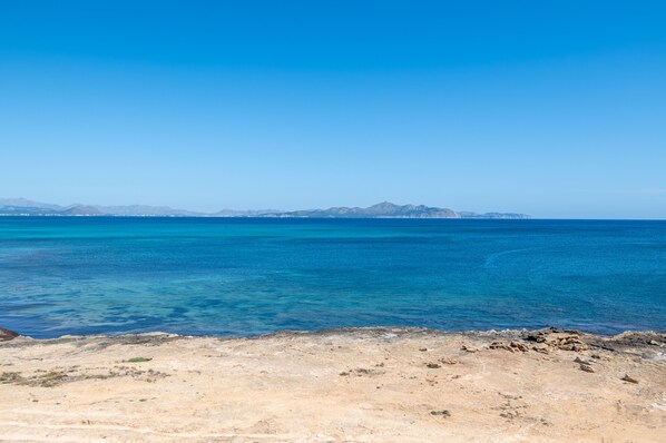 A coastal landscape with clear blue water, rocky shore, and distant mountains.