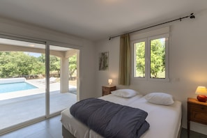 A bedroom with a bed, a nightstand, a lamp, a window, and a view of an outdoor swimming pool.