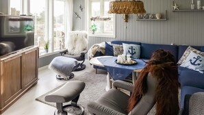 A cozy living room with a blue sofa, a grey armchair, and a television. There are decorative items like a hanging lamp and wall art. - Lovely home in Høvåg with WiFi (Høvåg)