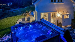 A hot tub with a stone surround, a patio with chairs, and a house with a lit interior. - Lovely home in Høvåg with WiFi (Høvåg)
