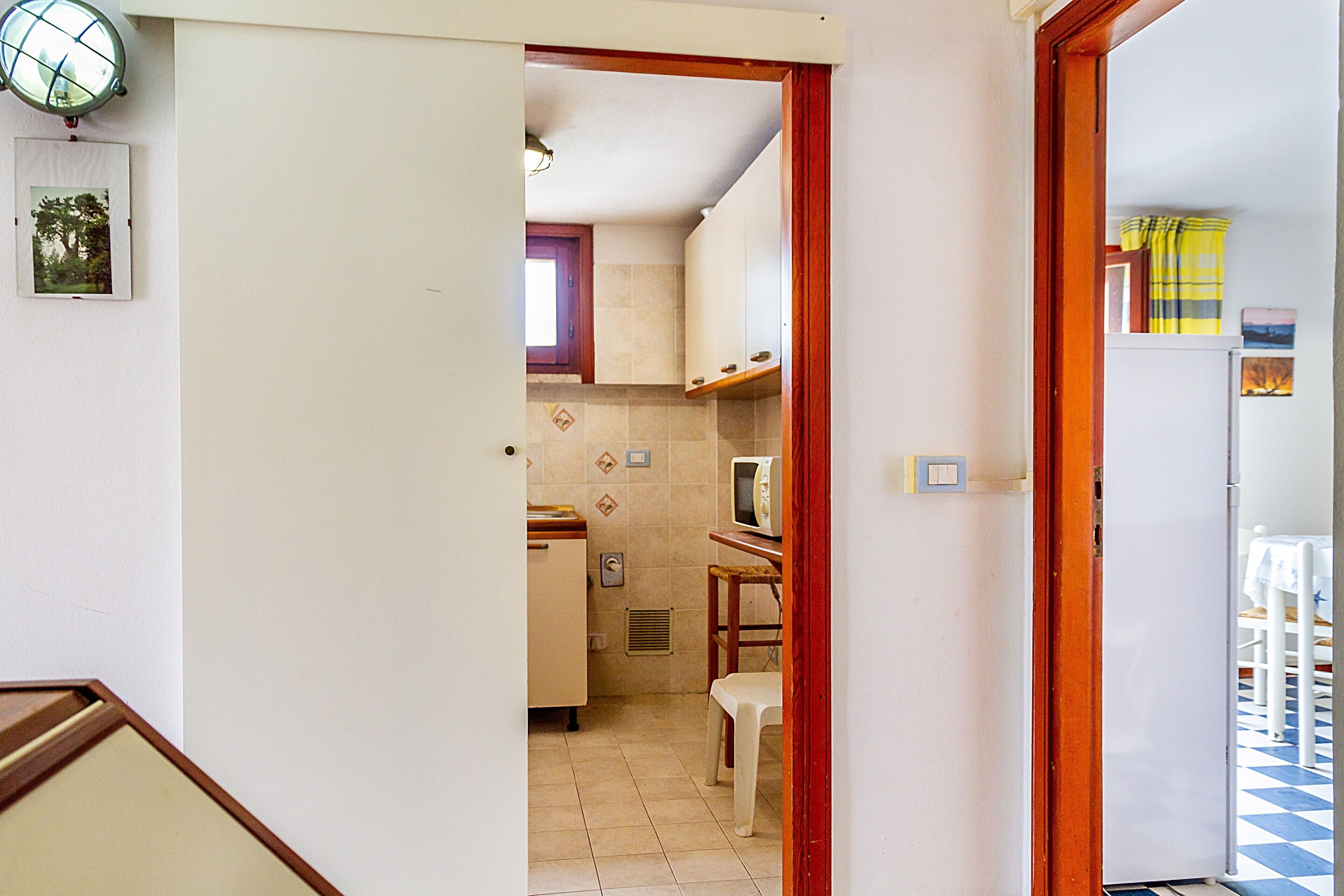 Ein schmaler Flur führt zu einer Küche mit Mikrowelle, Kühlschrank und einem Fenster mit Vorhängen.