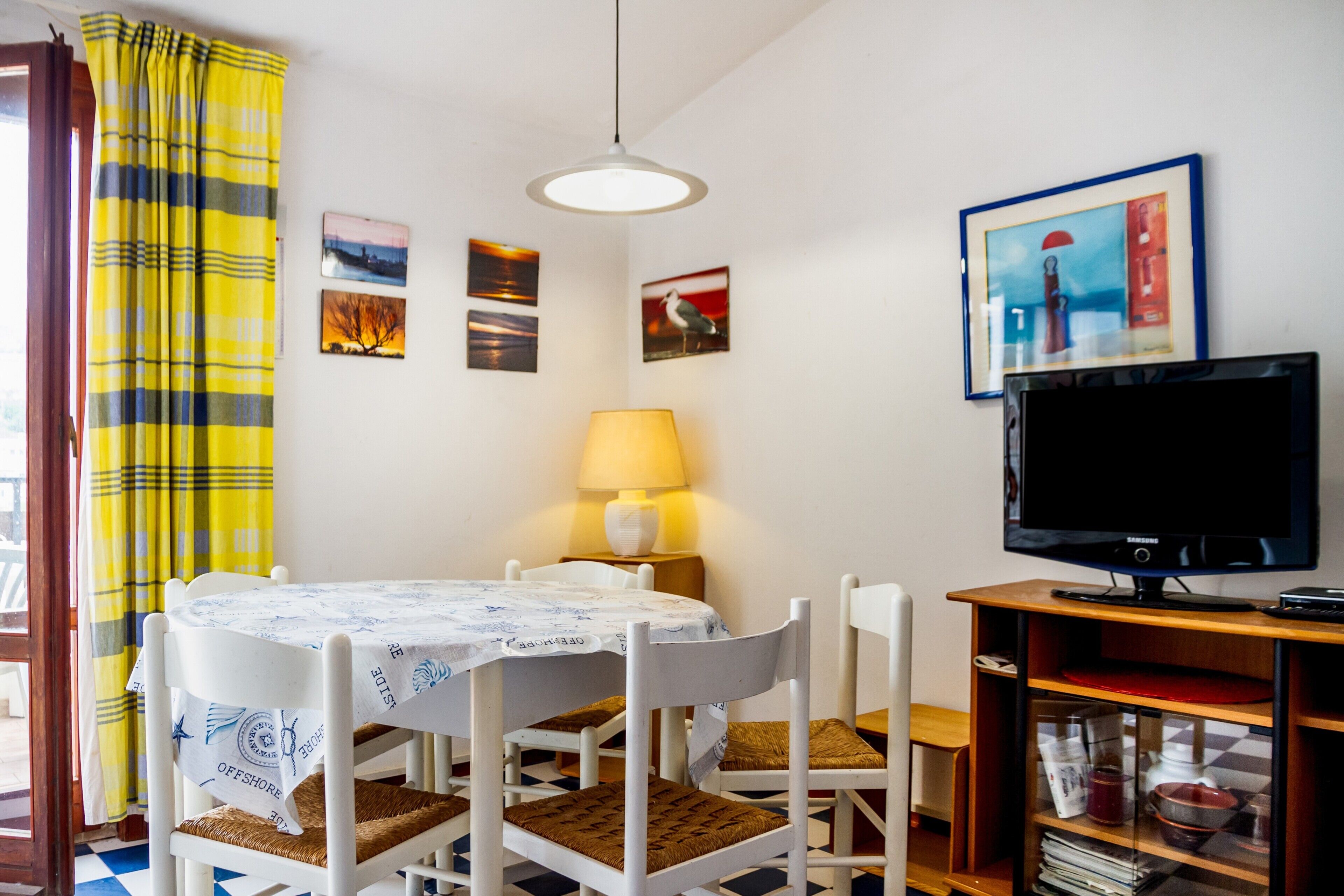 Et spiseområde med et bord, stole, en lampe, et fjernsyn og kunst på væggen.