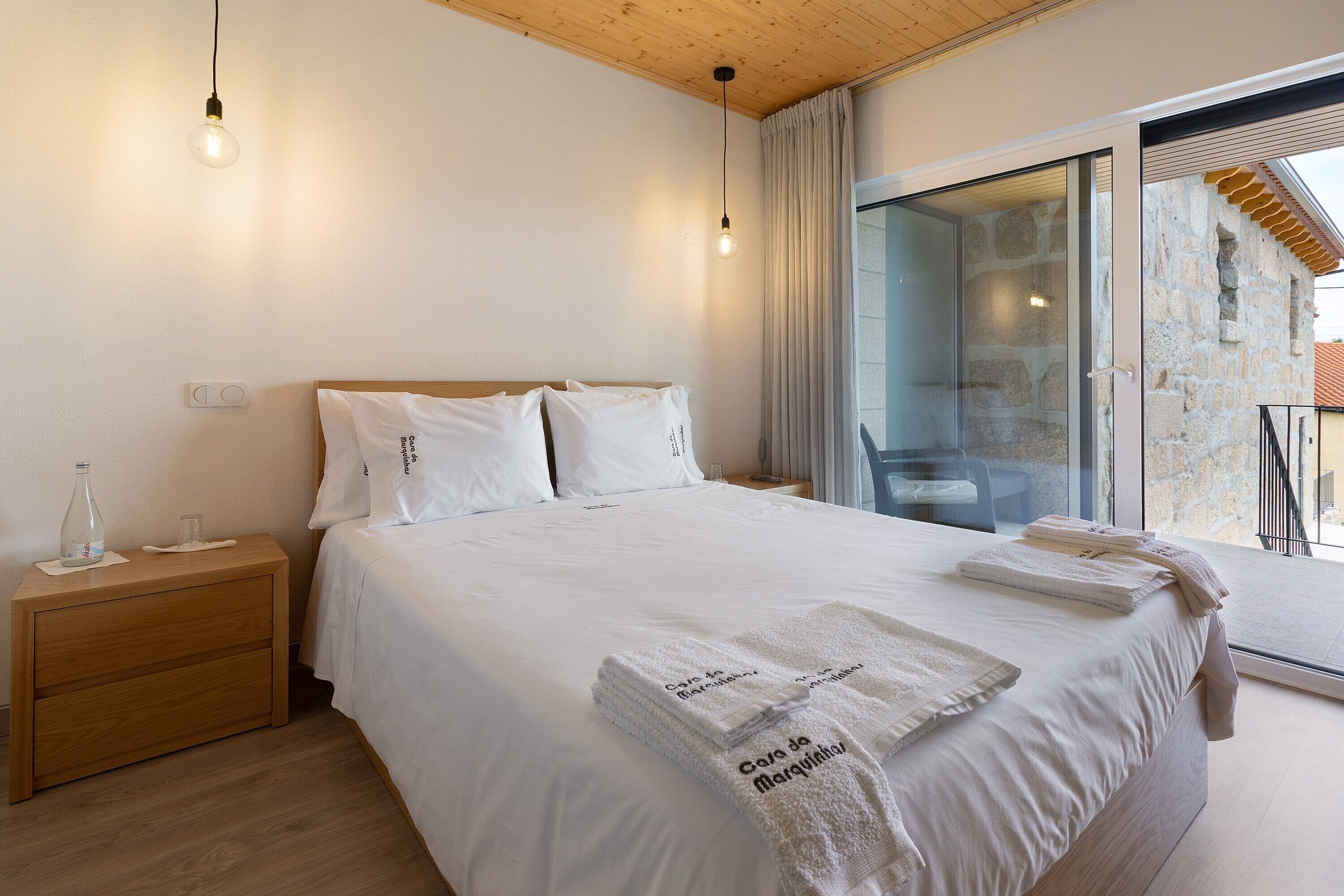 A bedroom with a wooden headboard, a nightstand with a bottle and glasses, a bed with white linens and towels, and a large sliding glass door leading to an outdoor area.