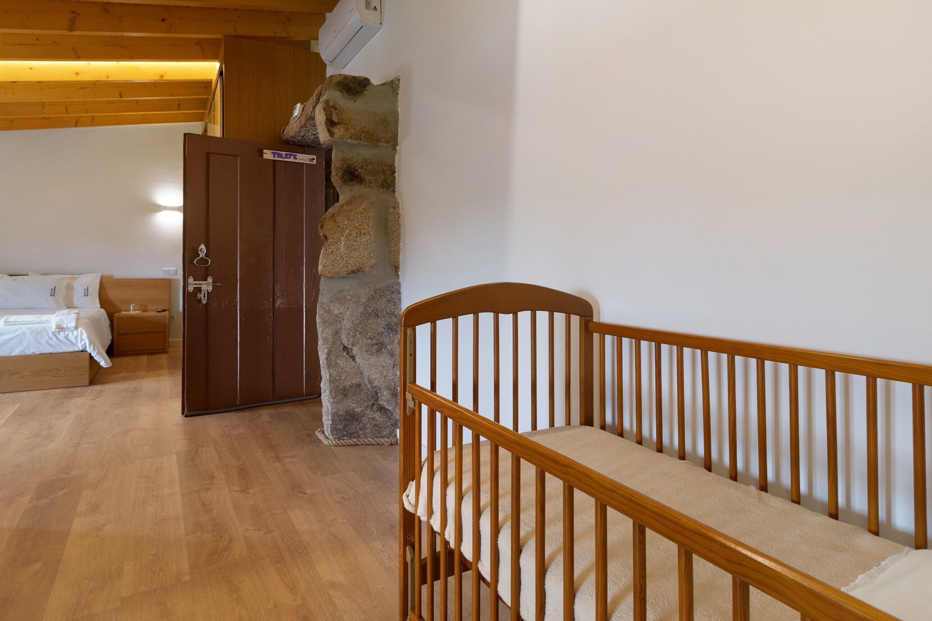Bressol de fusta en una habitació amb paret de pedra, una porta de fusta i un llit amb roba de llit blanca.