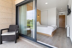 Une chambre d’hôtel moderne dotée d’une porte vitrée donnant sur un balcon, d’un lit avec du linge de lit blanc et d’une chaise en osier à l’extérieur.