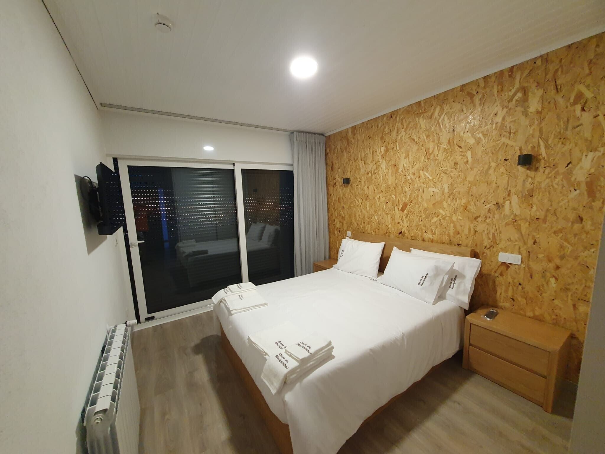 A modern hotel room with a large bed, a wooden accent wall, a sliding glass door to the balcony, and a built-in TV.