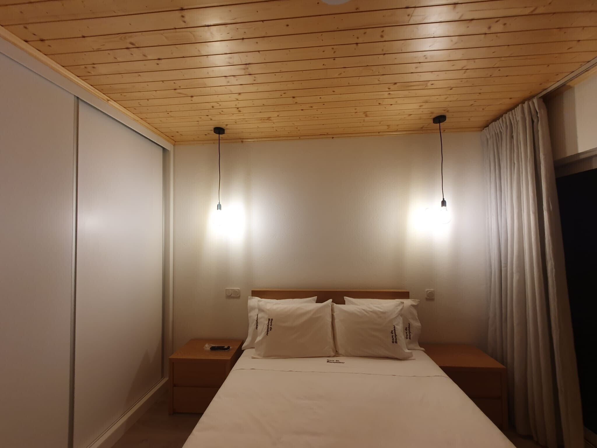 Un dormitorio con techo de madera, dos mesitas de noche, una cama con ropa de cama blanca y una cortina a la derecha.