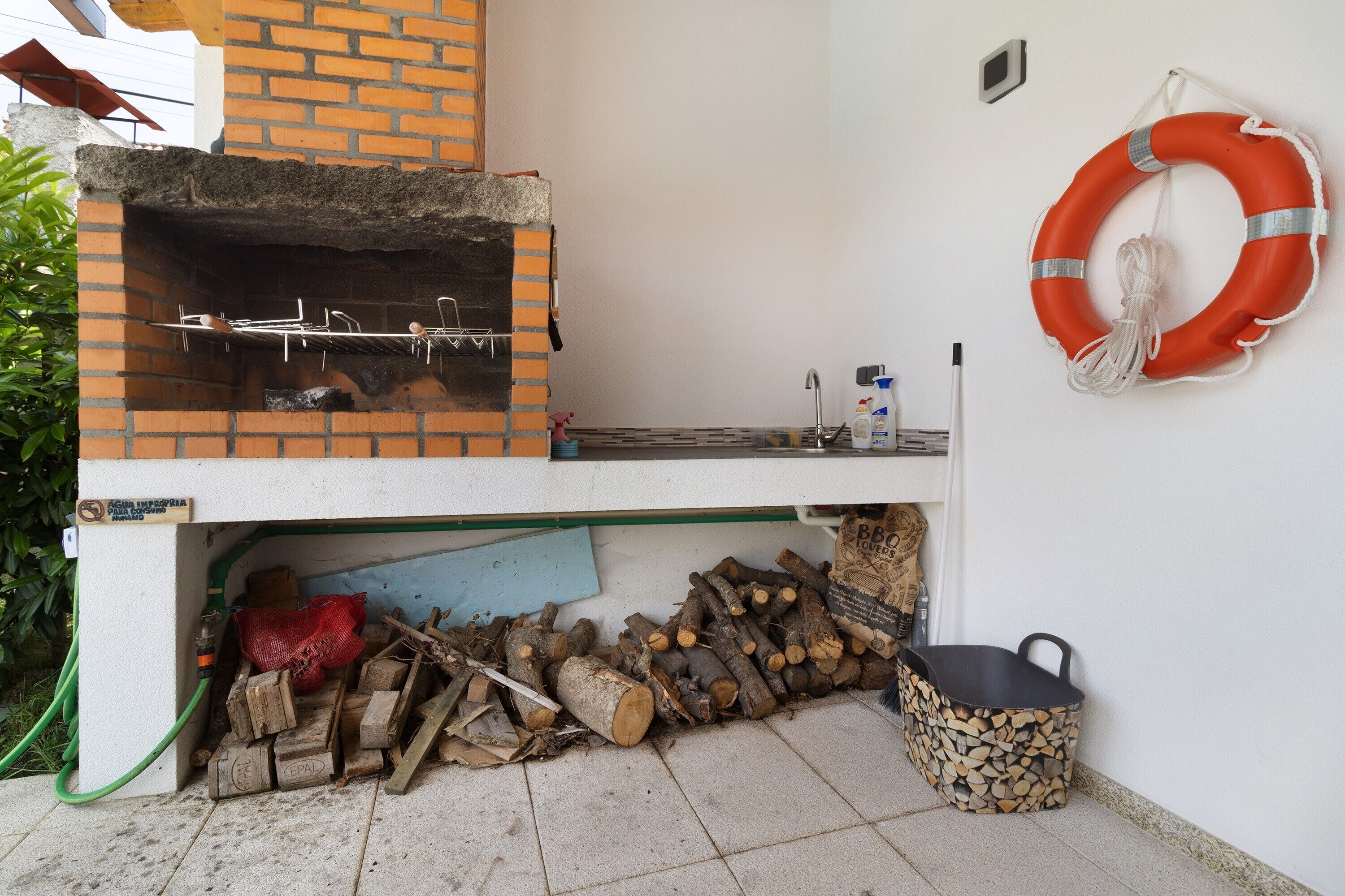 A brick-built outdoor grill with a metal rack, a life preserver on the wall, and a bucket of firewood.