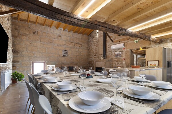 A stone-walled dining area with a long table set for a meal, wooden beams on the ceiling, and a fireplace in the background.