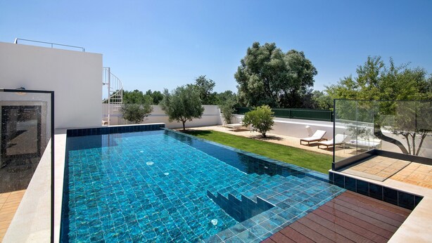 Una piscina rectangular con un borde de baldosas, rodeada por un edificio blanco y un área verde con árboles.