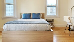 Ein Schlafzimmer mit einem Bett, zwei Fenstern, einer Klimaanlage, einem Sessel und einem kleinen Tisch.