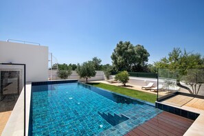 A rectangular swimming pool with a tiled deck, surrounded by a white building and a grassy area with trees. - Apartment 'Monte Gerações 3' with Shared Pool, Wi-Fi and Air Conditioning (Almancil)