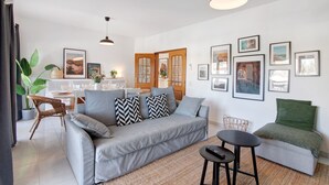 A modern living room with a grey sofa, a small round table, and a green chair. There are framed pictures on the wall and a dining area in the background.