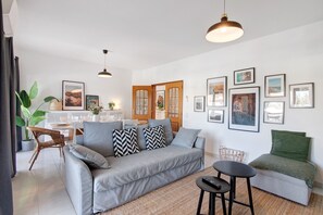 Una sala de estar moderna con un sofá gris, una mesita redonda y un sillón verde. Hay cuadros enmarcados en la pared y un comedor al fondo.