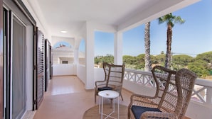 A balcony with wicker chairs, a small table, and a view of greenery and buildings.