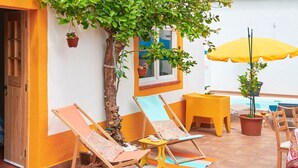 Um pátio com uma árvore, duas cadeiras de descanso, um guarda-sol amarelo e uma pequena mesa.