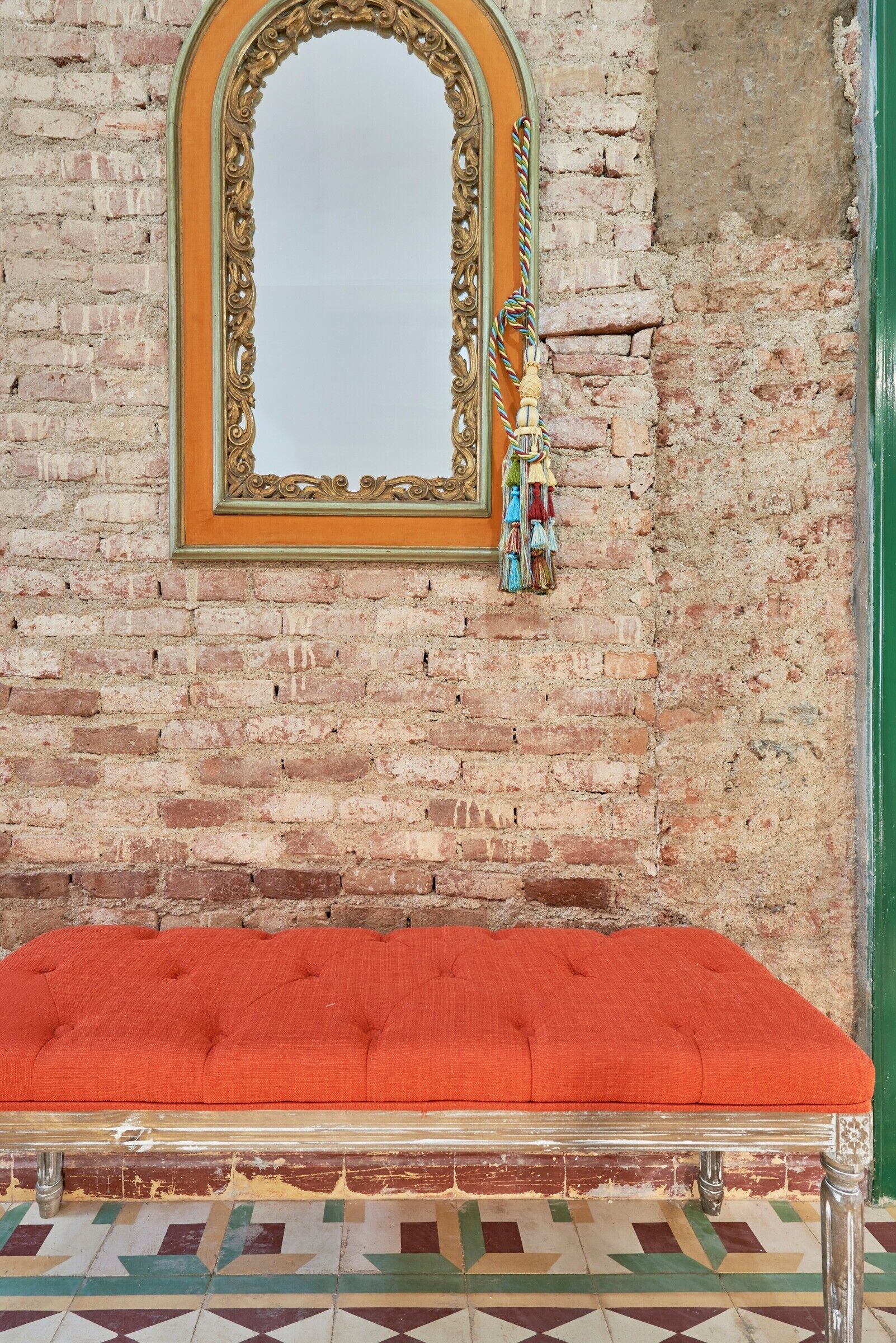 A room with a brick wall, a mirror with an ornate frame, a red cushioned bench, and a patterned floor.