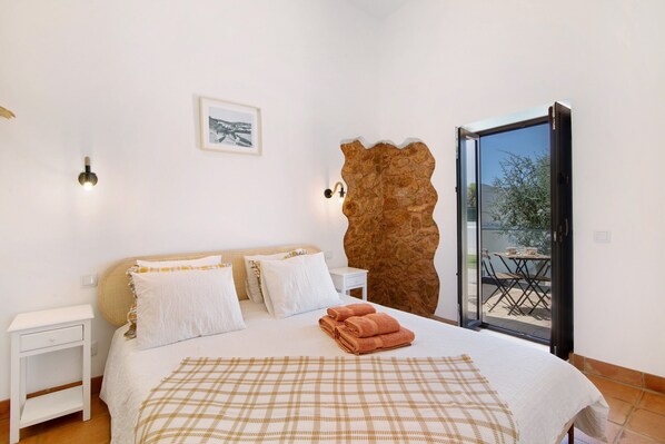 A bedroom with a bed, white nightstands, a large window with a view, and a stone wall.