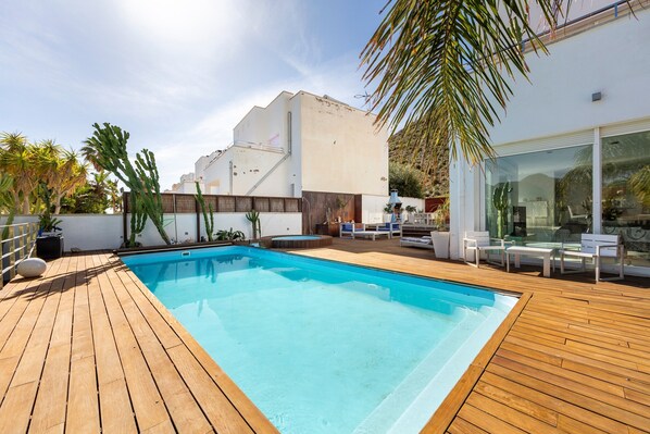 Une piscine dotée d’une terrasse en bois, entourée de palmiers et bordée d’un bâtiment moderne aux grandes fenêtres.