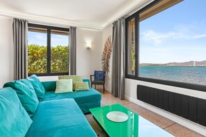 A modern living room with a teal sofa, a green coffee table, and a large window offering a view of the sea and distant hills. - Exception Maison Beachfront Holiday Home with Private Garden and Wi-Fi (Fréjus)