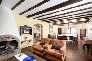 A living room with a fireplace, patterned sofa, dining area, and a piano. - Country House 'Cal Eloi Janet' with Mountain Views, Private Pool and Wi-Fi (Sant Marti Sarroca)