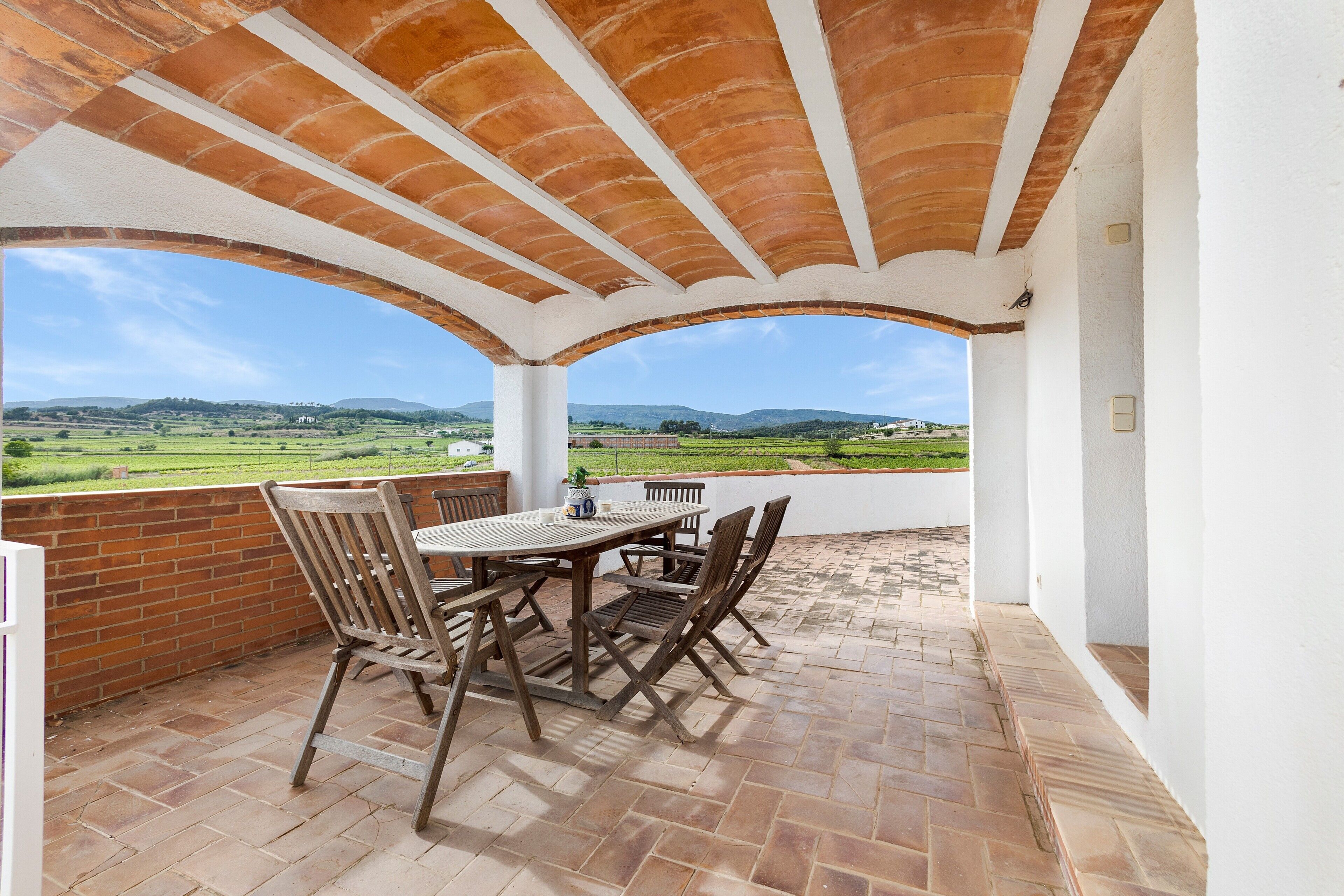 Un patio cubierto con una mesa y sillas, un muro de ladrillo y vistas a campos verdes y colinas.
