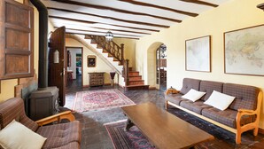 A cozy living room with a fireplace, a sofa, a wooden table, and a staircase leading to another room. - Country House 'Cal Eloi Janet' with Mountain Views, Private Pool and Wi-Fi (Sant Marti Sarroca)
