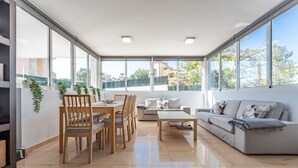 A spacious living area with a dining table, chairs, a sofa, and a coffee table. Large windows provide a view of the outside.