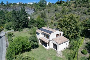 A two-story house with a solar panel roof, surrounded by trees and a hillside.