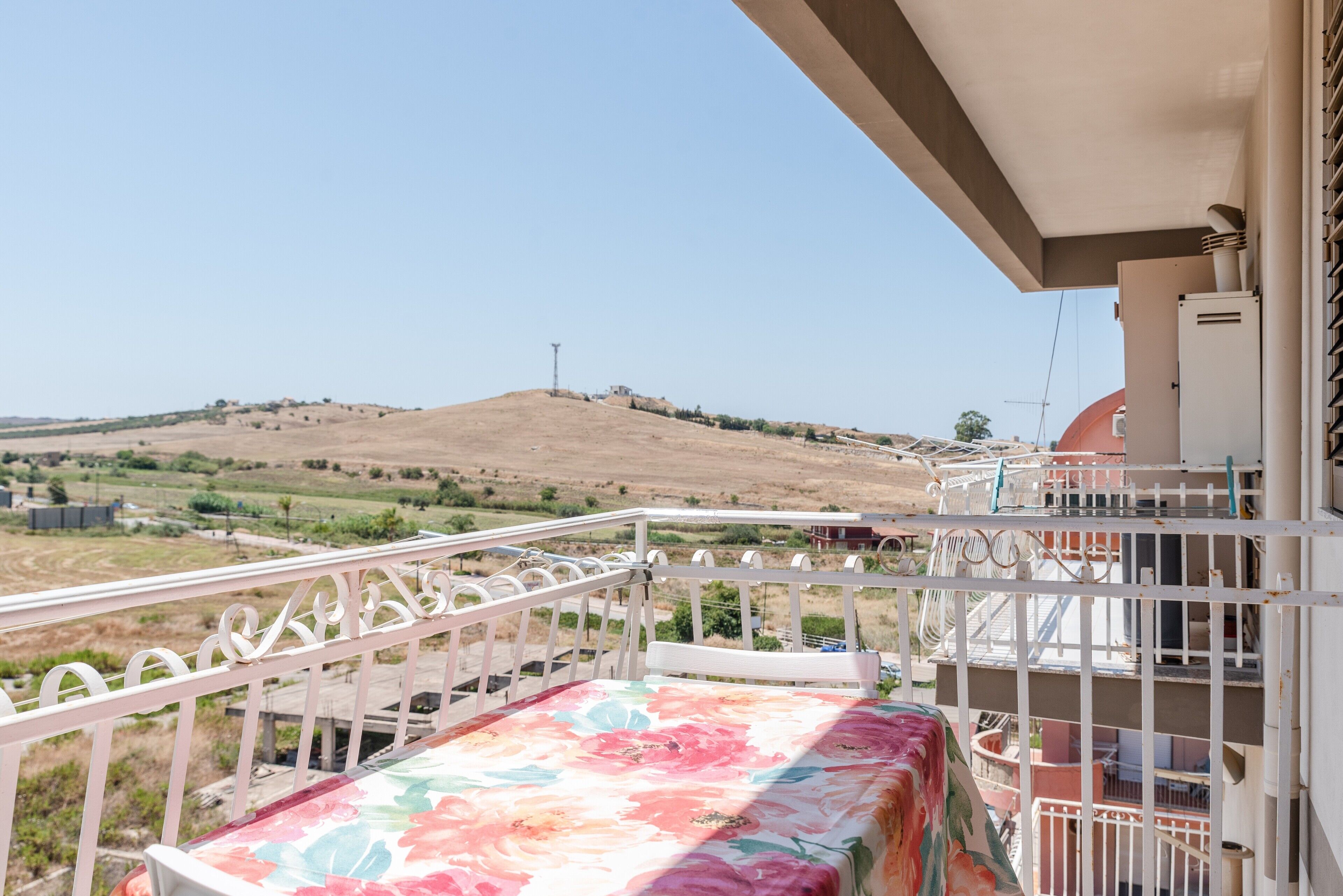 A balcony with a floral tablecloth, a white railing, and a view of a hilly landscape.